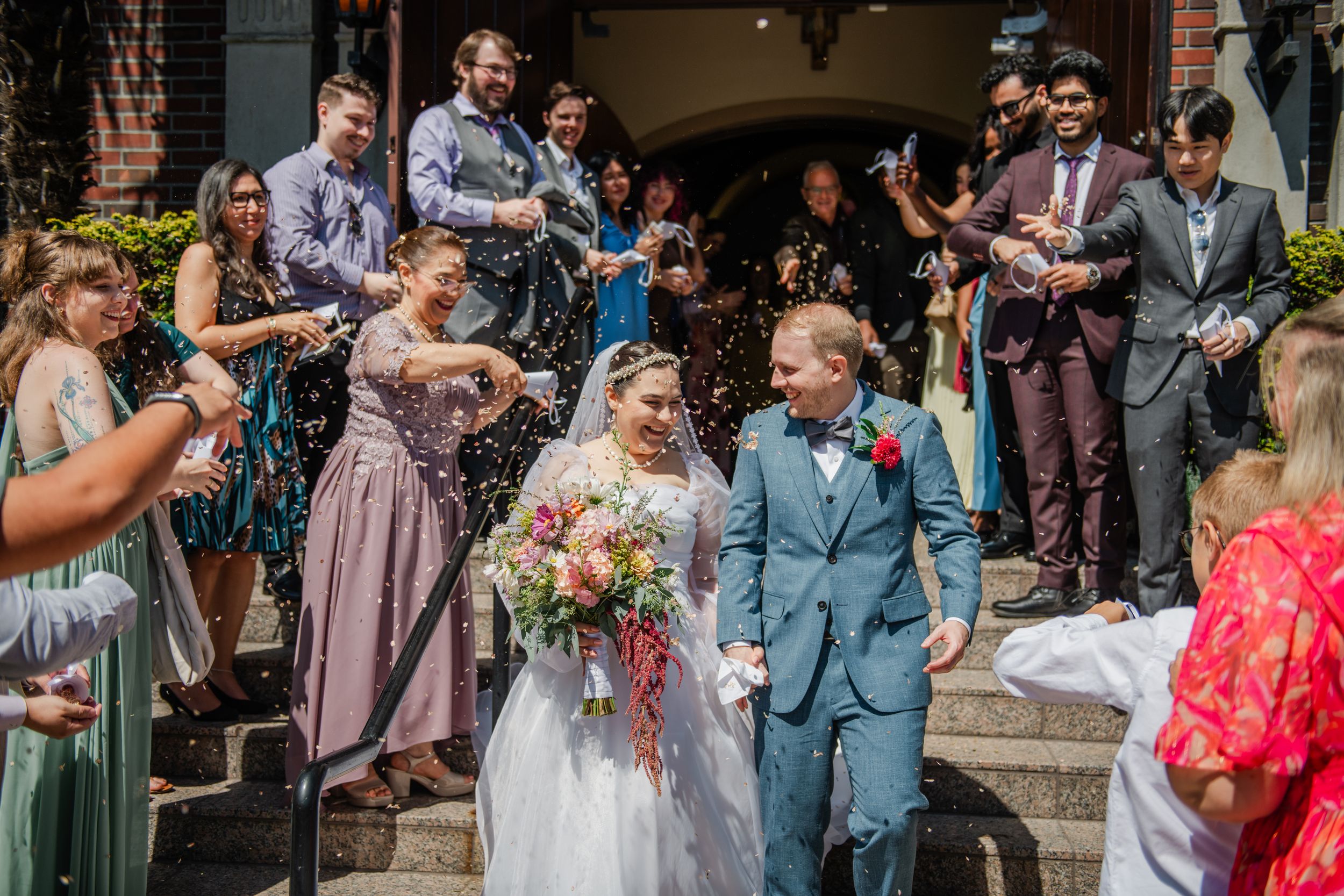 Newlyweds walk through confetti shower as guests celebrate outside church steps after wedding ceremony.