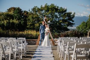 Couple stands at end of white aisle runner with empty chairs and mountain backdrop.