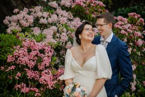 A couple shares a joyful moment in front of blooming pink rhododendrons in a garden setting.