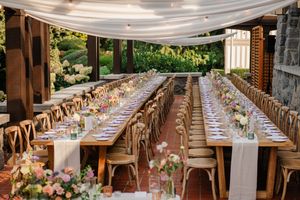 Elegant outdoor wedding reception setup with long wooden tables, cross-back chairs and white draping overhead.