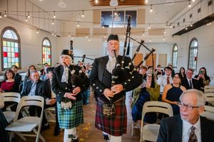 Bagpipers in kilts lead a wedding processional through an elegant white venue with string lights.