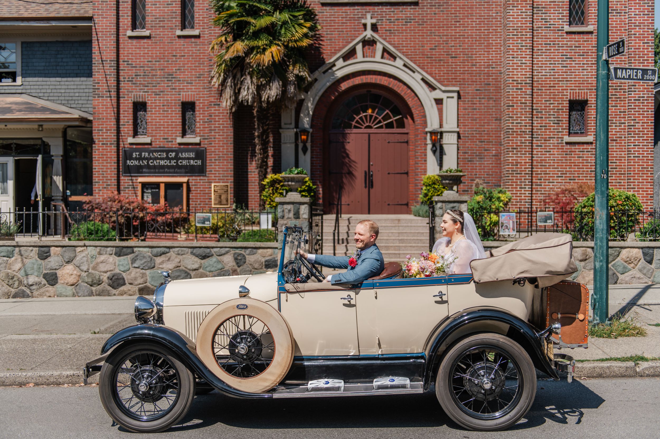 Couple riding in a vintage cream-colored convertible antique car in front of a historic brick church building.