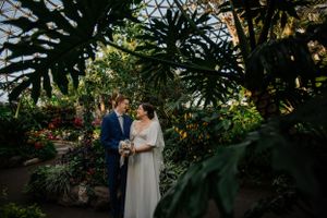A couple stands among tropical plants and flowers in a garden conservatory setting.