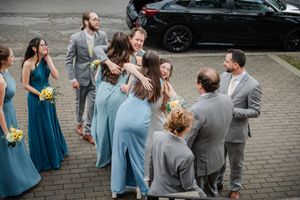 Wedding party in blue dresses and gray suits sharing joyful hugs outdoors near a parking lot.
