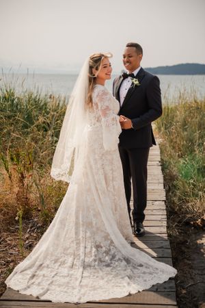 A couple poses on a wooden boardwalk with ocean views, showcasing a detailed lace wedding gown.
