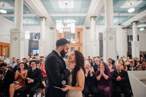A couple shares their first kiss at their wedding ceremony in a grand hall while guests applaud in celebration.