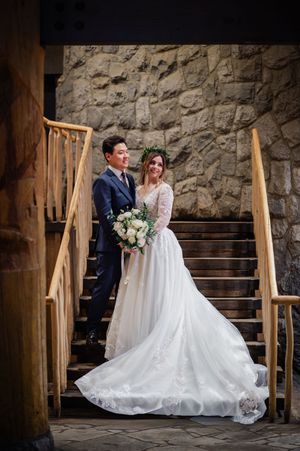Newlywed couple sits together on rustic wooden steps against a stone wall in an elegant wedding setting.