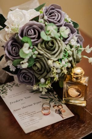Wedding details with purple and white rose bouquet, perfume bottle, and invitation on wooden surface.