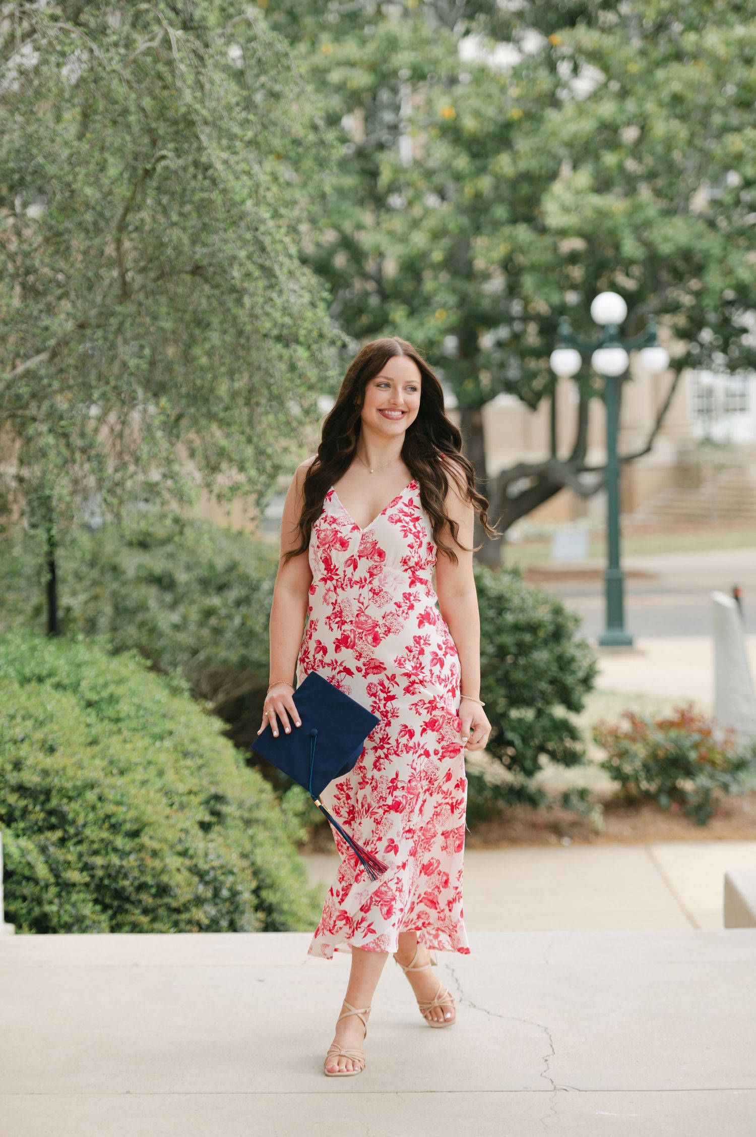 A smiling woman walks in a red floral dress holding a navy diploma cover outdoors.