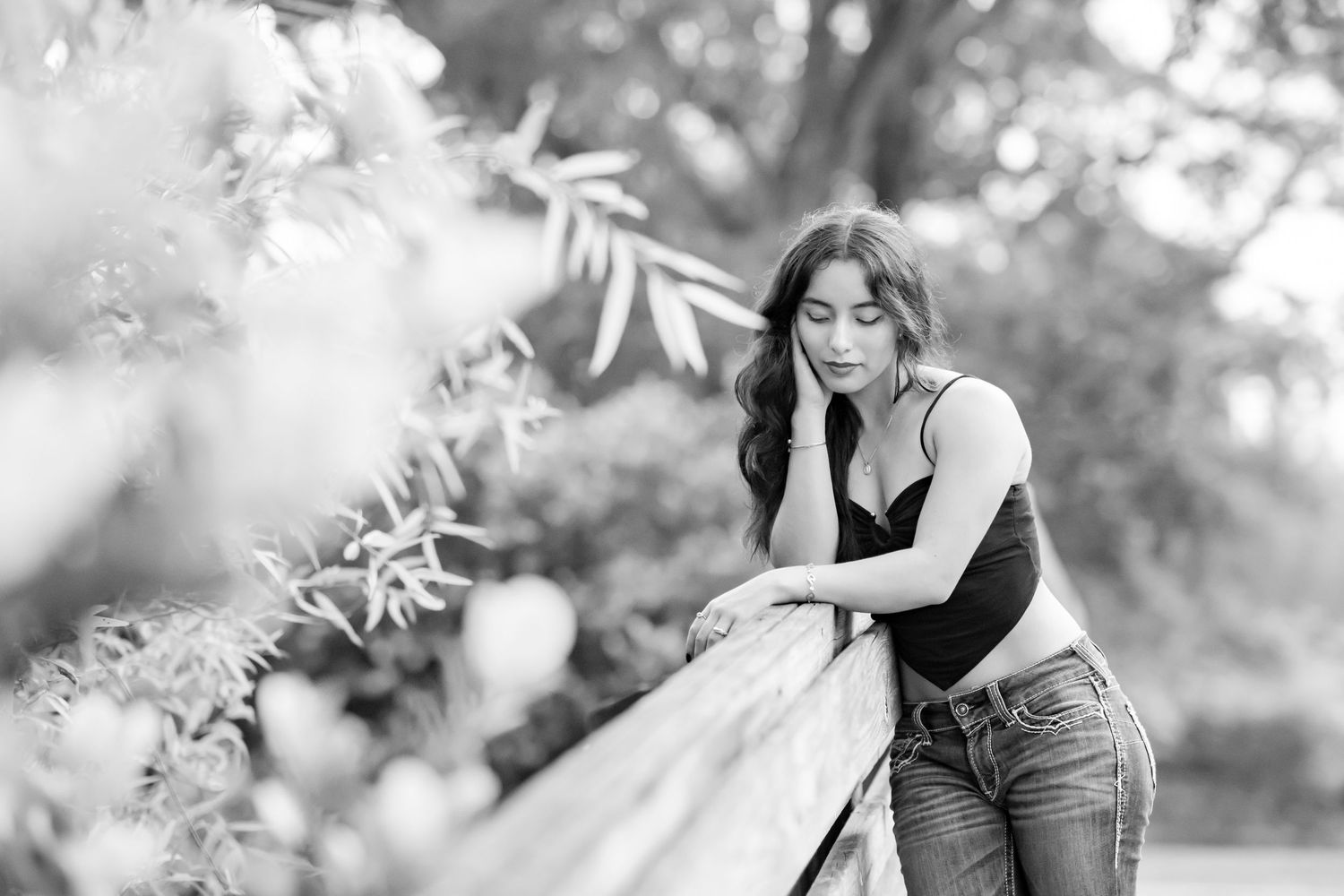 Black and white outdoor portrait showing figure leaning contemplatively against fence in dreamy natural garden setting.