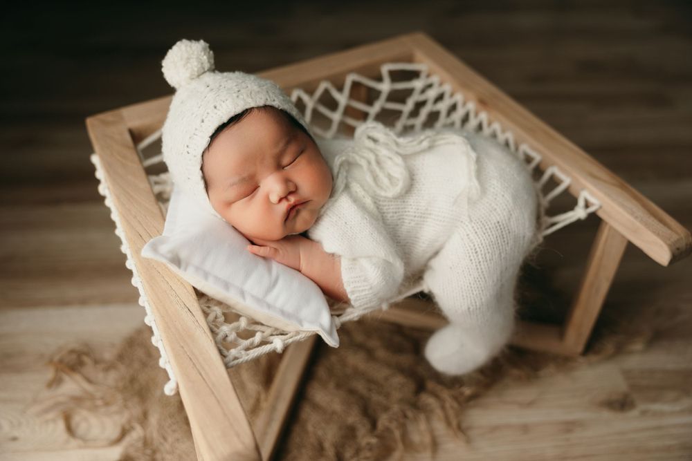 A sleeping baby in white knitwear rests on a small wooden bed prop.