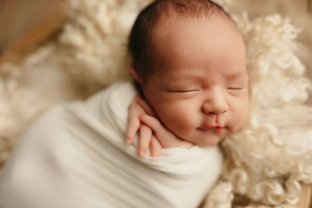 A peaceful newborn sleeping while swaddled in white against a cream background.