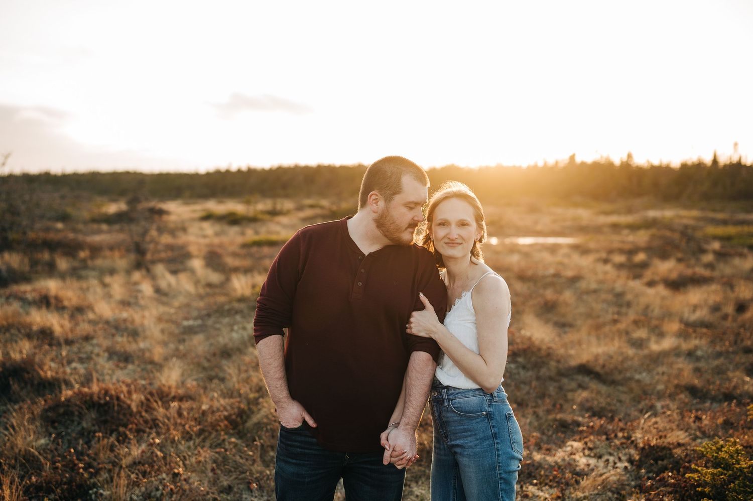 engagement session with a woman hugging the arm of her husband to be, a large woodsy field behind them.