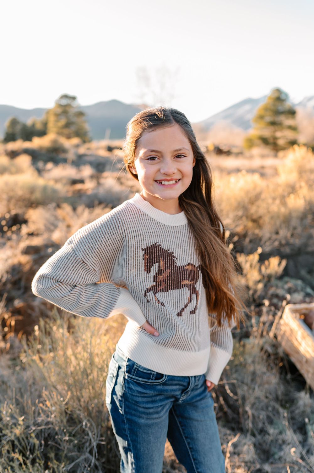 A young person in a horse-patterned sweater and jeans poses outdoors against a mountain landscape at sunset.