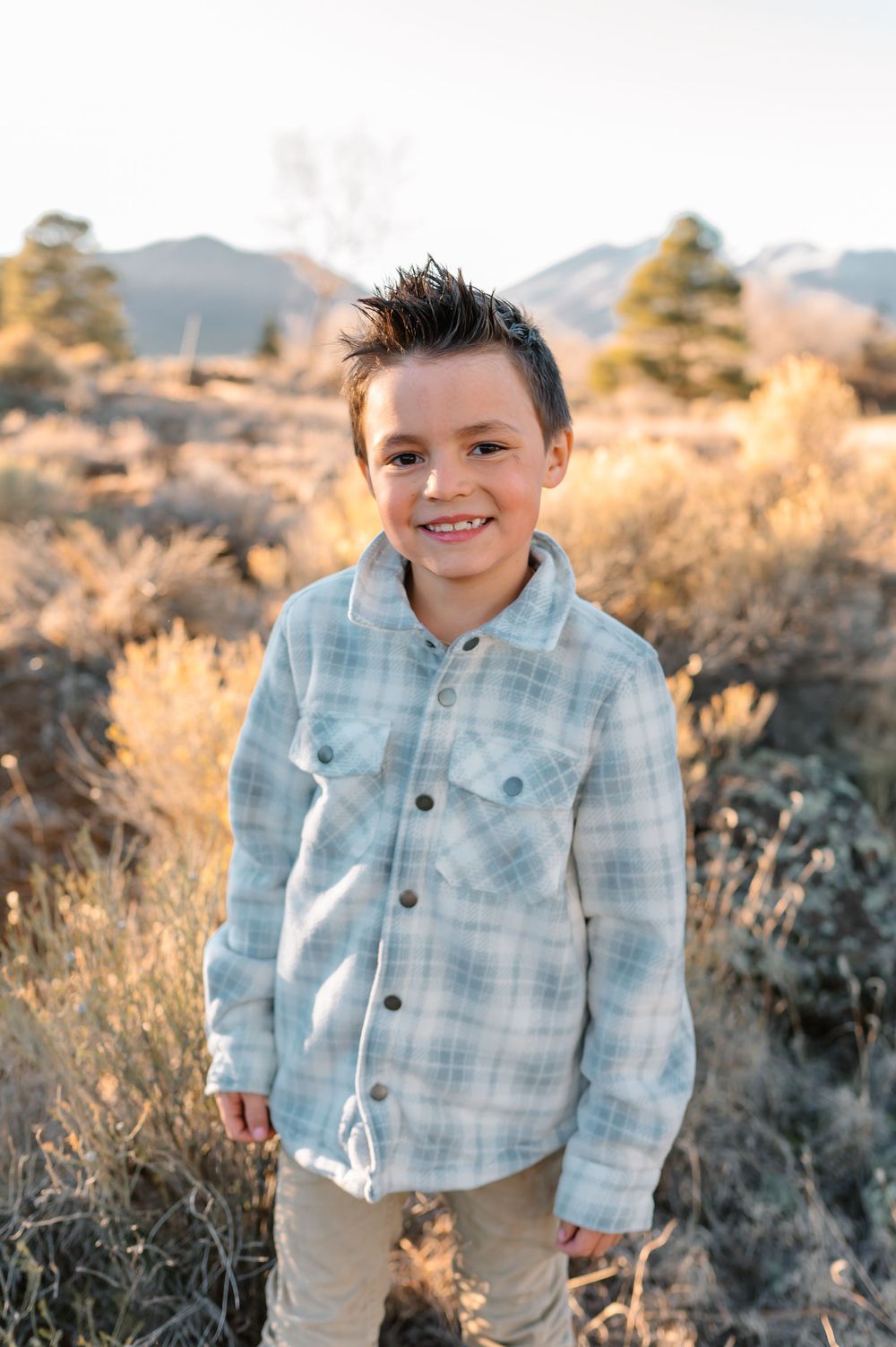 A child in a light blue plaid shirt stands in a desert landscape with sagebrush and mountains during golden hour.