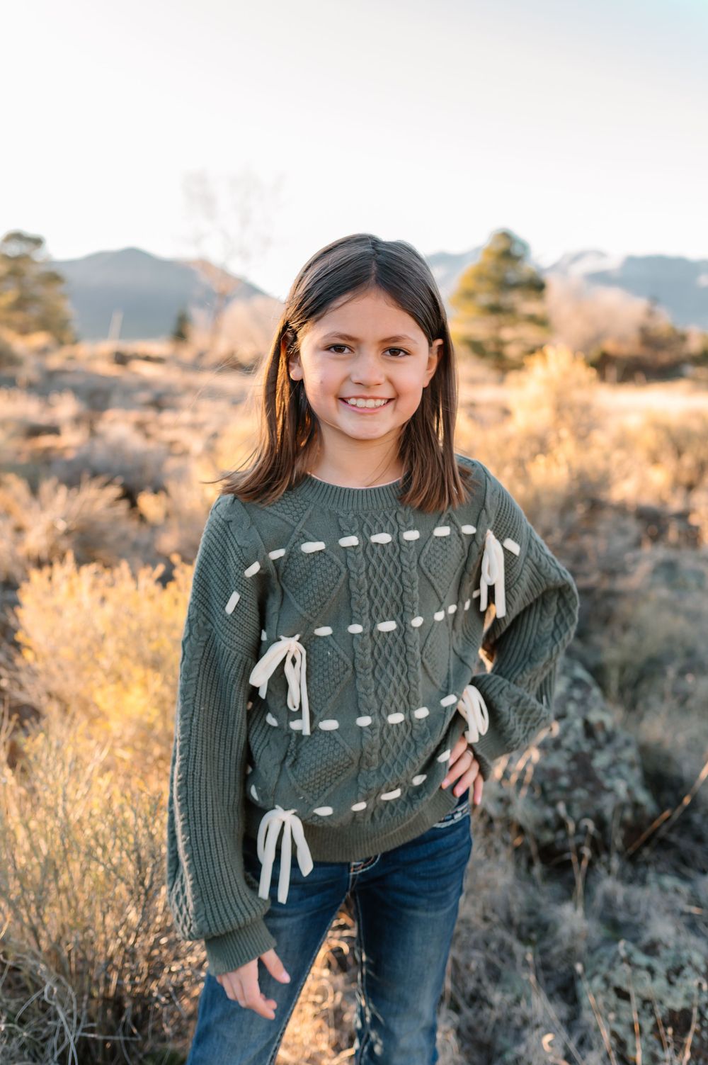A person in a green knit sweater with ribbon details stands outdoors in a desert landscape at sunset.
