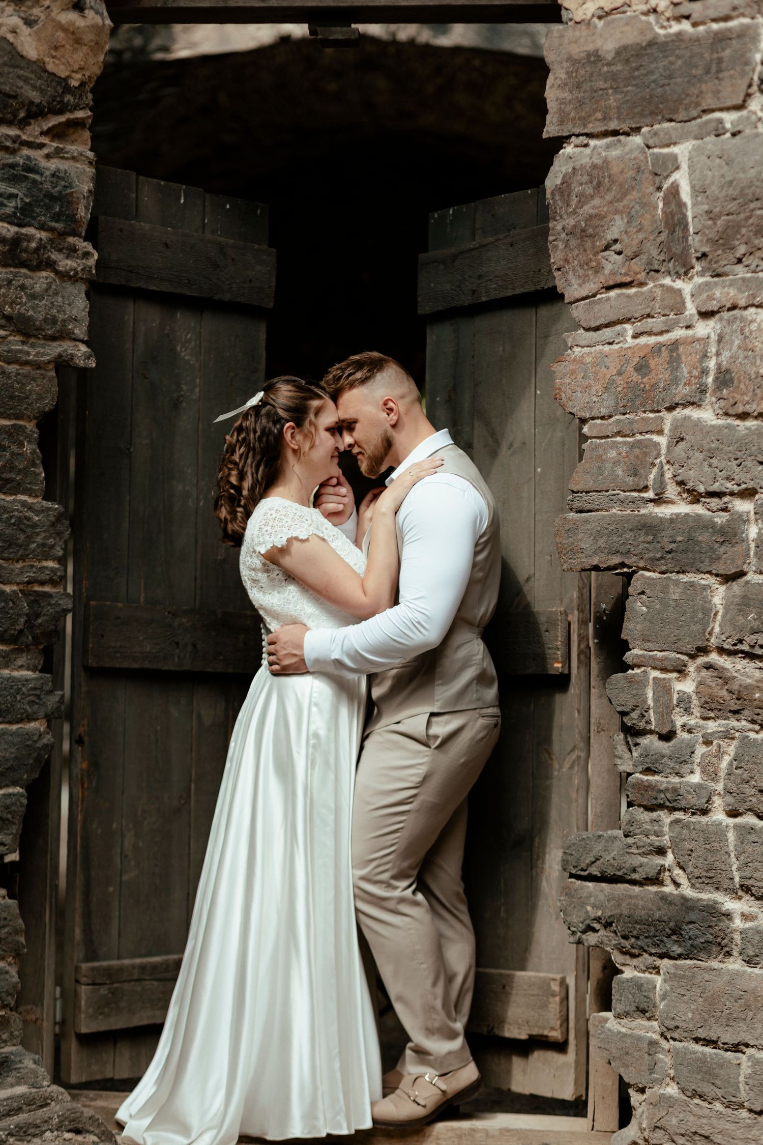 Bride and groom embracing in front of a rustic wooden door set in a stone wall.