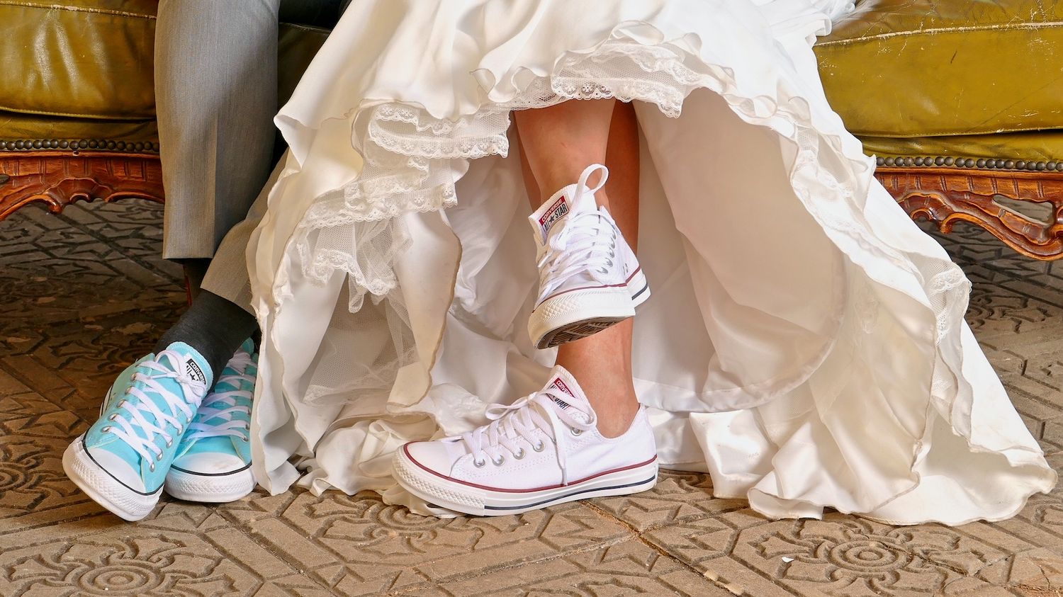 Dois pares de ténis Converse, um branco e outro azul, surgem por baixo de um vestido de noiva branco.