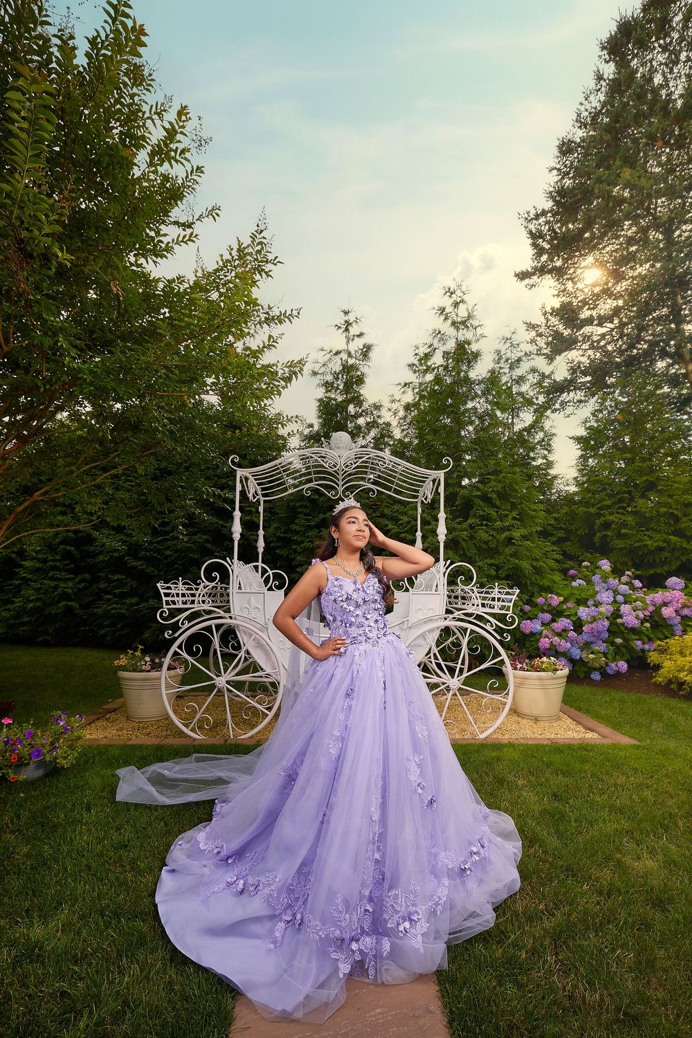 Young woman in lavender quinceañera ball gown with floral details poses by white carriage in lush garden.