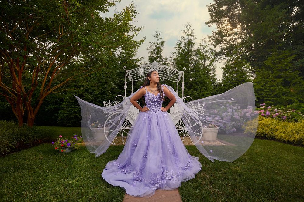 Young woman in lavender quinceañera ball gown posing before a white carriage in a lush garden.