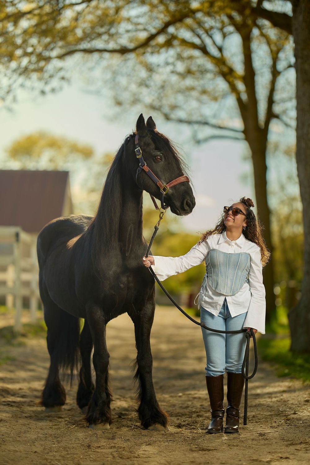 Woman in white blouse and jeans leading a black horse on a sunny tree-lined path at a ranch.