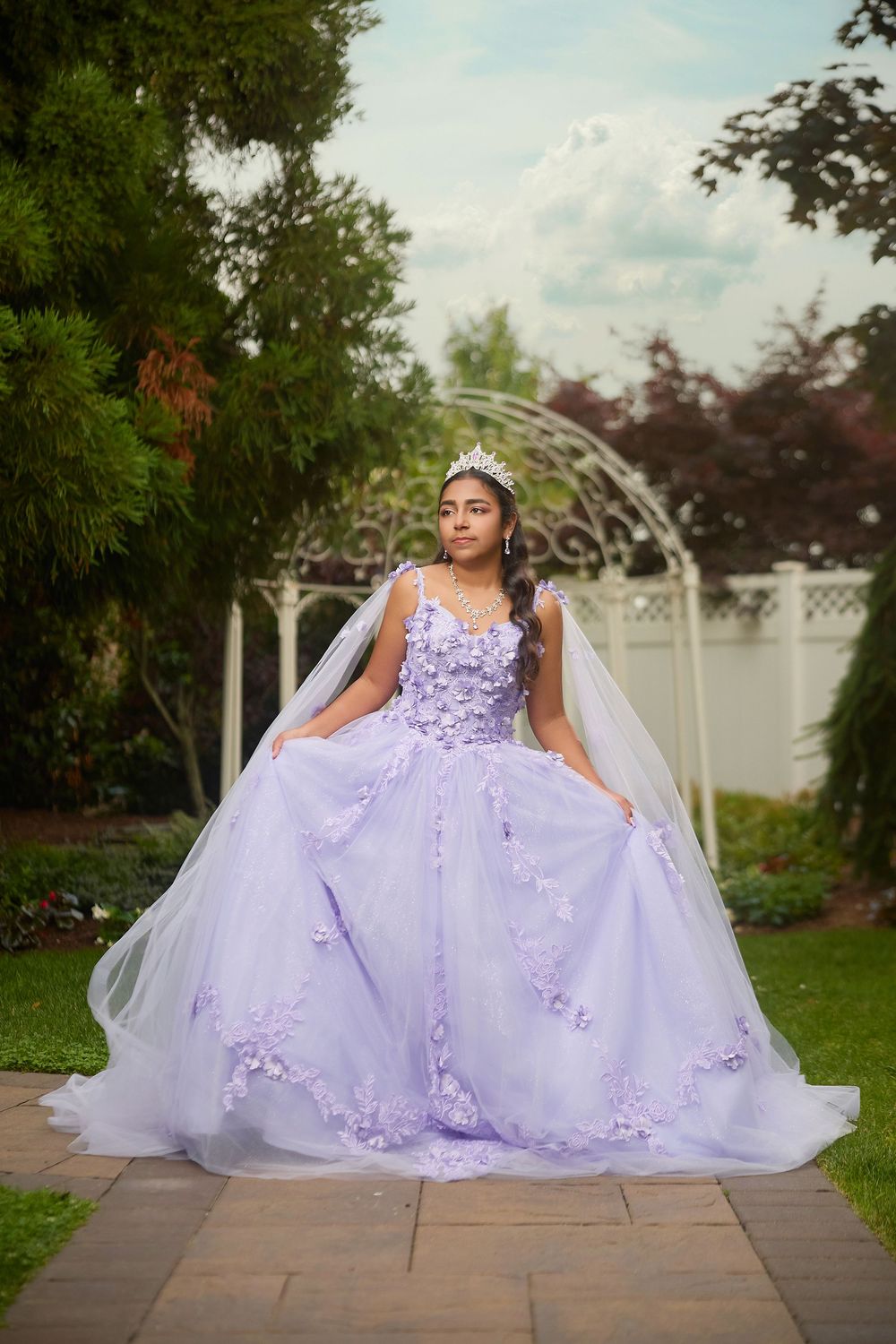 Teen girl in lavender quinceañera ball gown with floral details and tiara in garden.