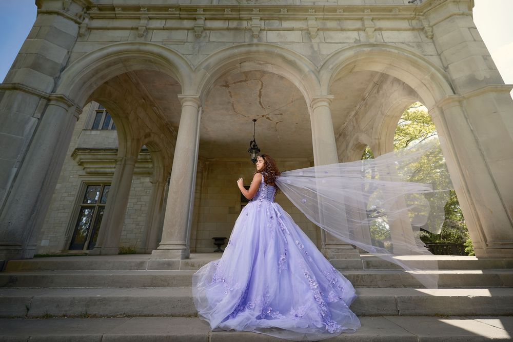Young woman in lavender ball gown with flowing veil poses gracefully on stone steps of elegant classical building.