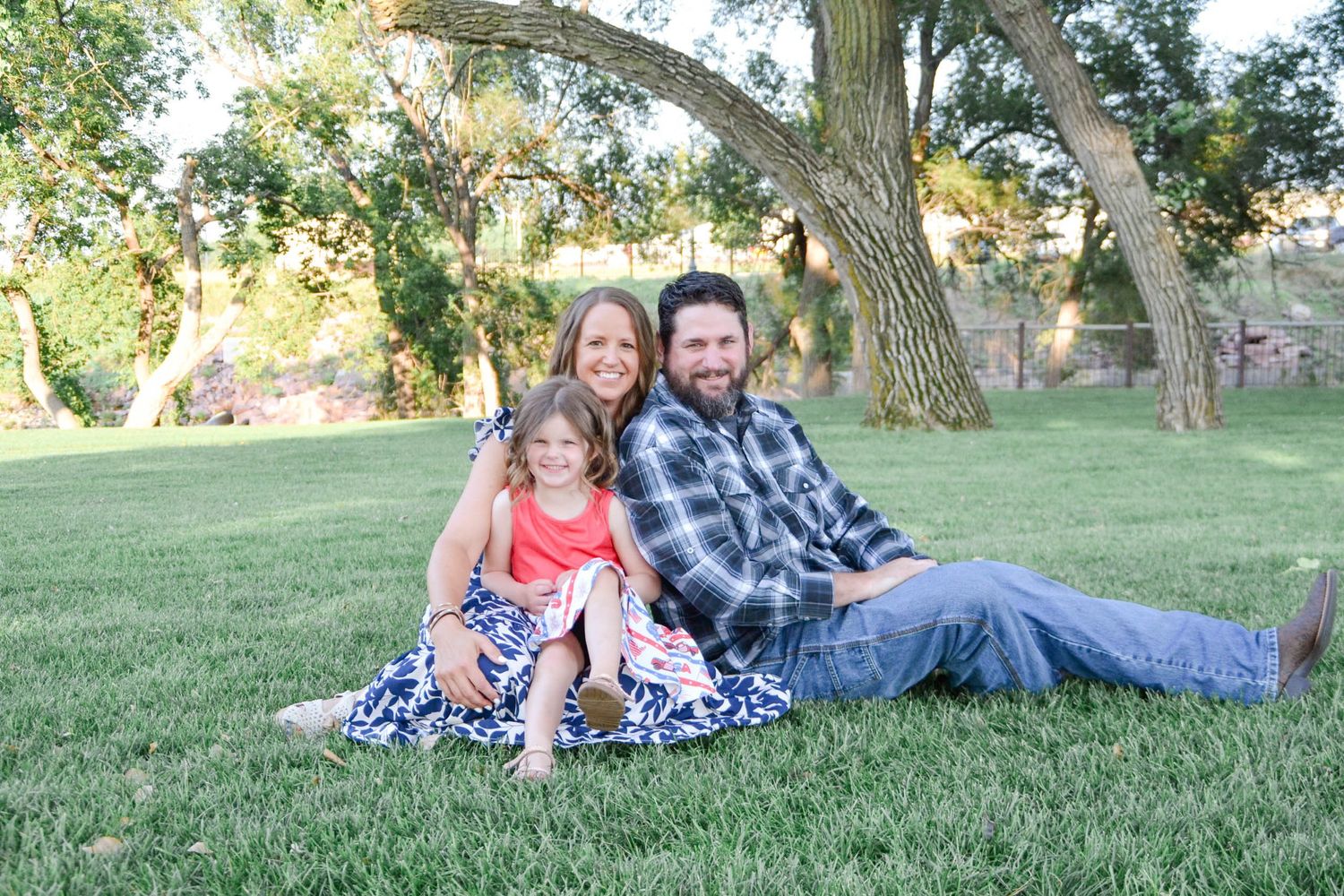 A small group relaxes together on a grassy lawn under the shade of large trees on a sunny afternoon.
