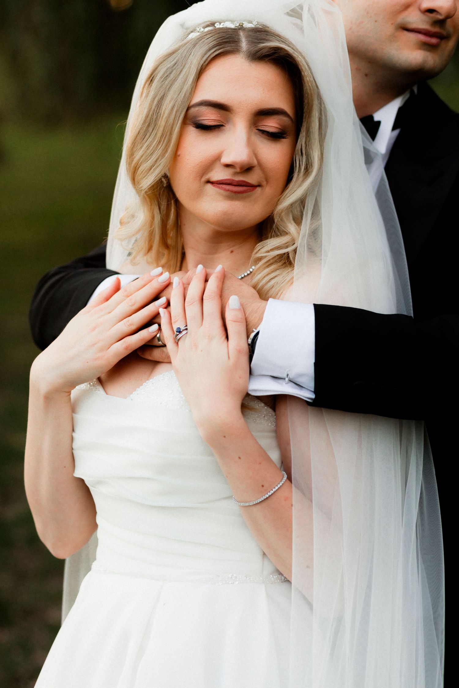 Bride and groom embracing in golden sunset light among trees, wedding dress train flowing on green grass.