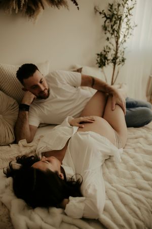 A couple in white clothing laying intimately together on white bedding in a moody, romantic setting.