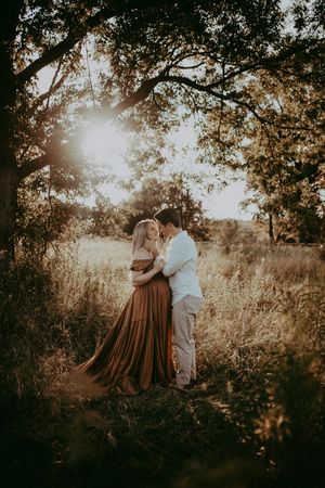 A couple embraces in a romantic outdoor setting during golden hour as sunlight filters through tree branches.