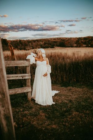 A vintage christening gown hangs on a wooden fence in a rustic field at sunset.
