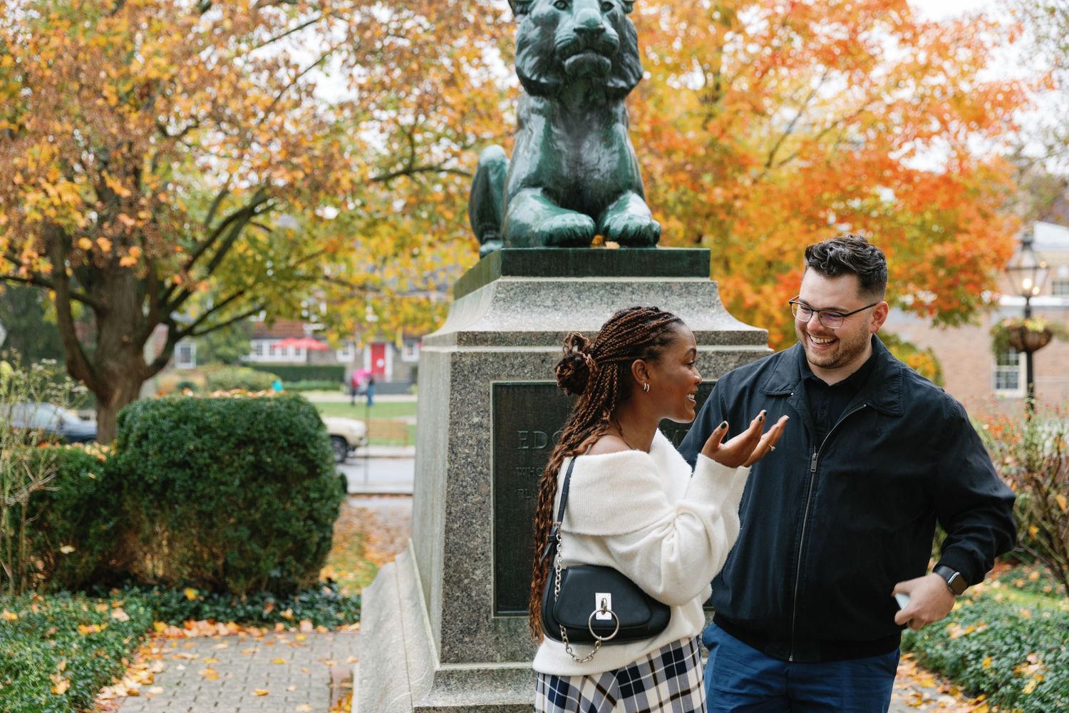 Two people share a candid moment laughing together by a statue in a park during autumn with colorful fall foliage.