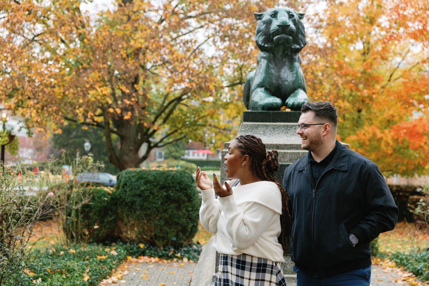 A couple shares a romantic moment under fall foliage near a sculptural bust in a park setting.