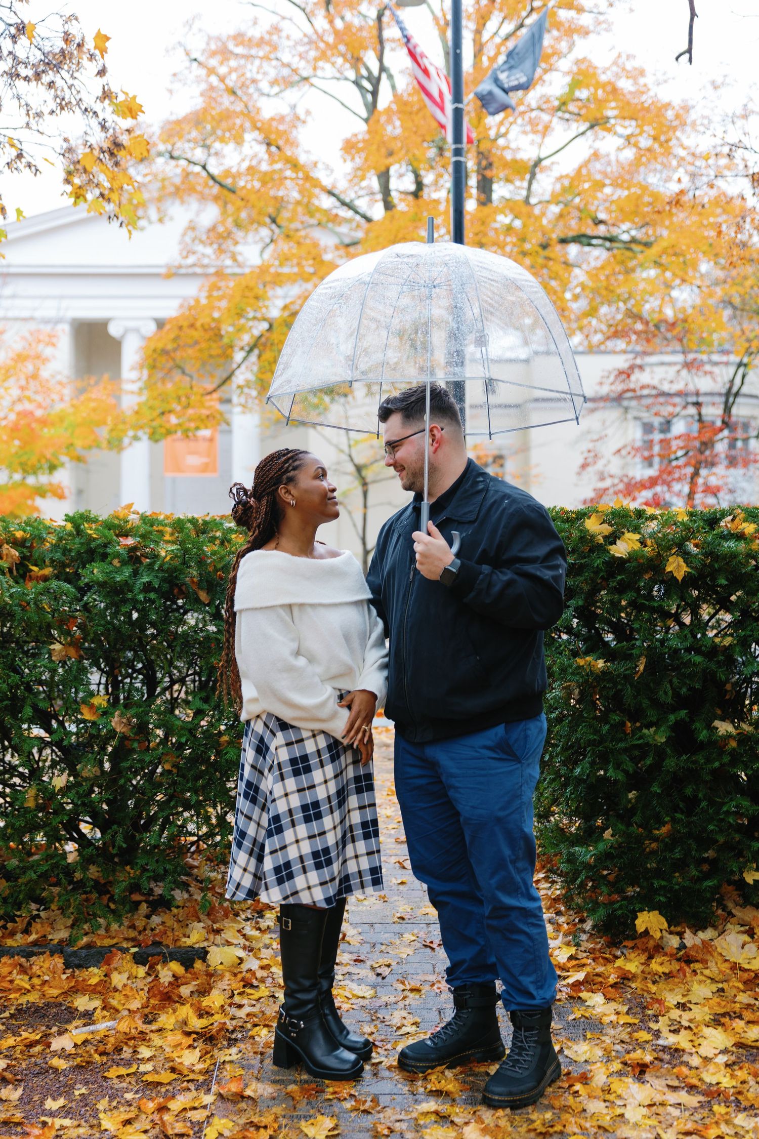 Two people share a clear bubble umbrella on an autumn day with golden leaves and a white building in the background.
