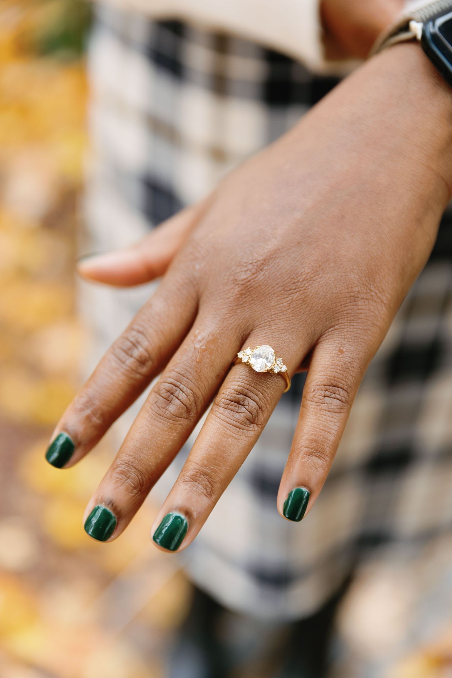 A hand wearing a sparkling diamond engagement ring and forest green nail polish against a blurred plaid background.