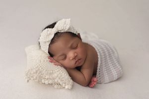Newborn baby girl sleeping on a creamy white blanket, her head and hand on a tiny cream lace pillow. She is wearing a matching lace headband
