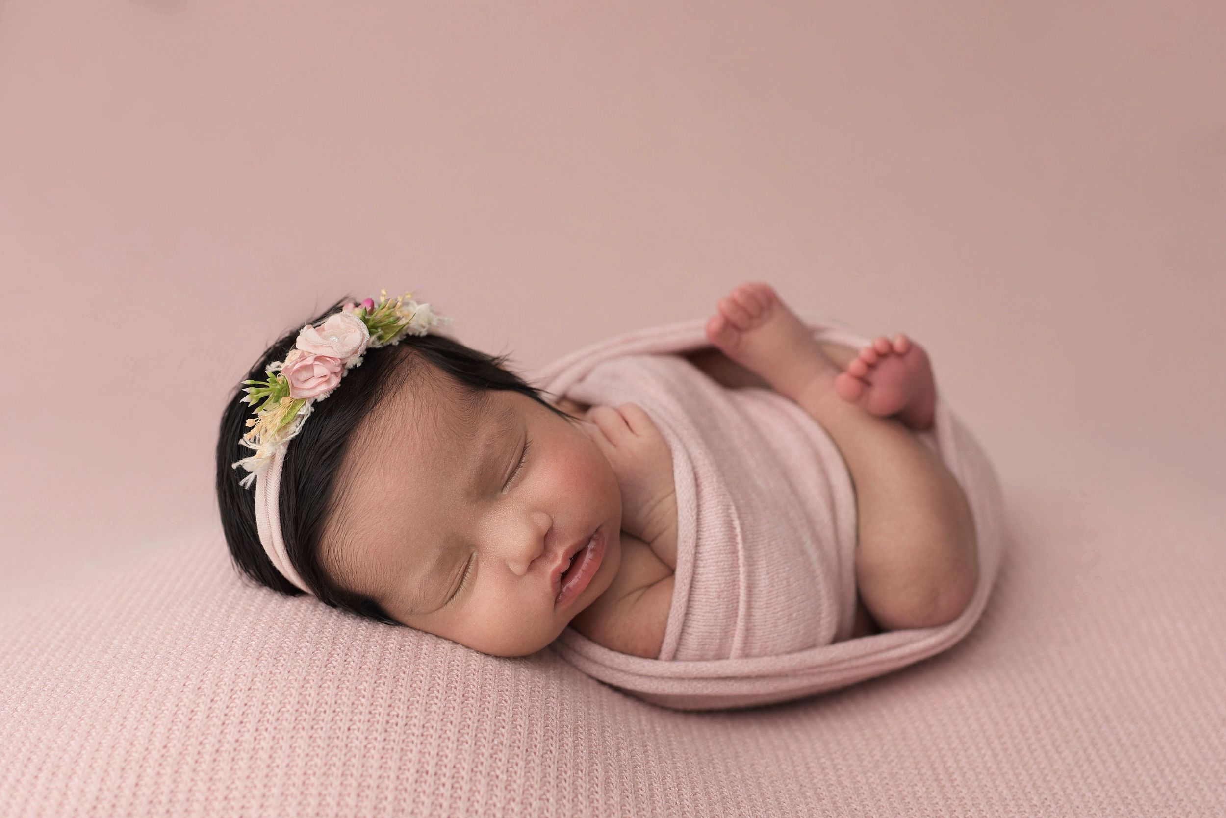 Newborn girl sleeping on her back in a snug light pink wrap on a matching light pink blanket.