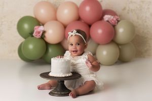 Baby girl with her birthday cake on a stand, the background is a simple small balloon arch in shades of greens and pinks.