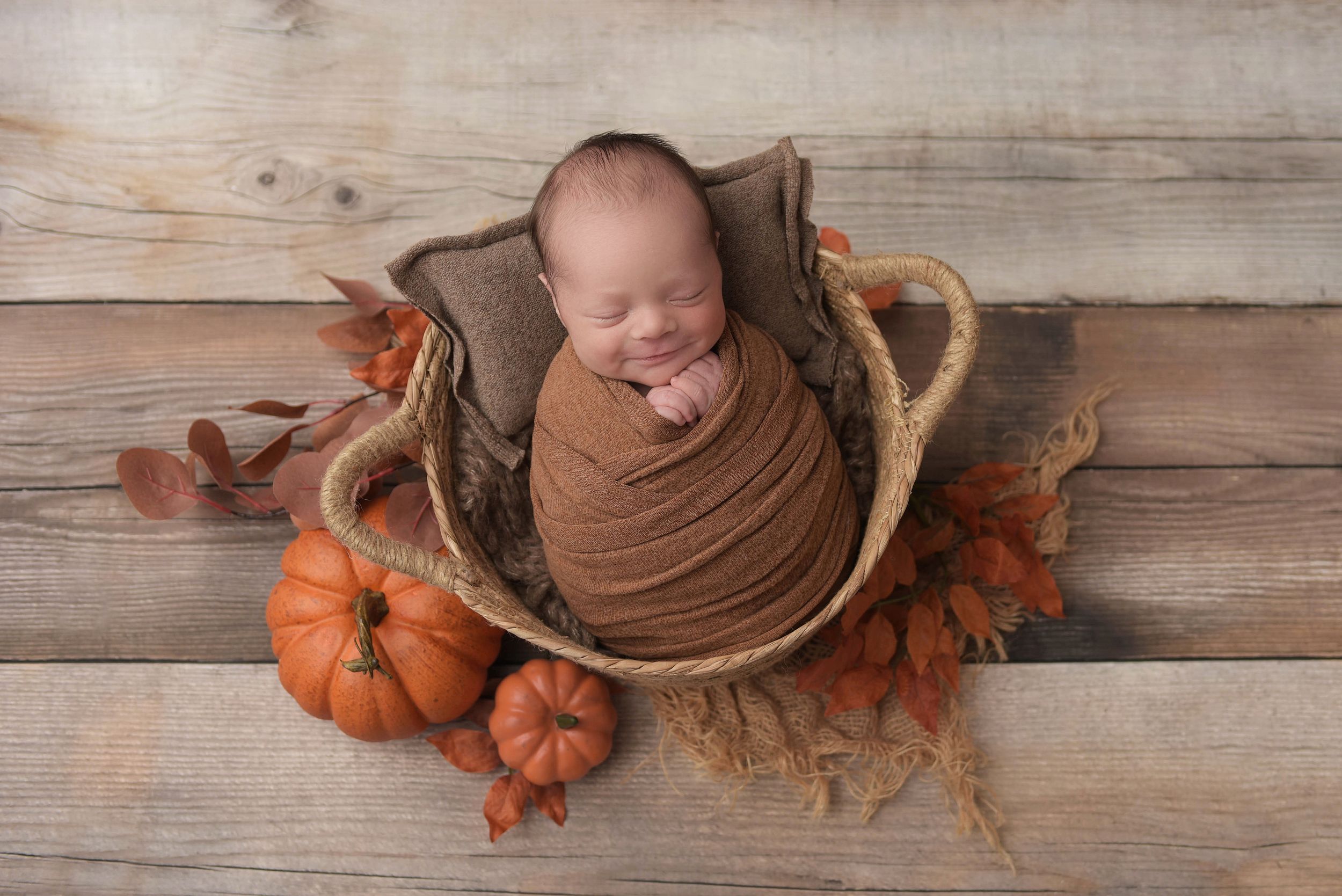 Newborn boy smiling in his sleep, in a brown swaddle in a rustic basket surrounded by orange pumkins and fall leaves.
