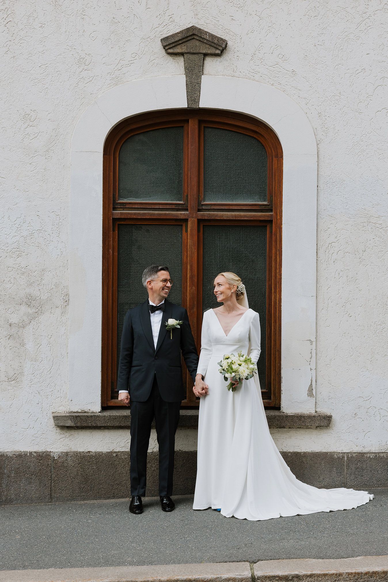 Brudepar som står ganske rett mot kamera, hånd i hånd, ser på hverandre. De innrammes av et stort vindu.