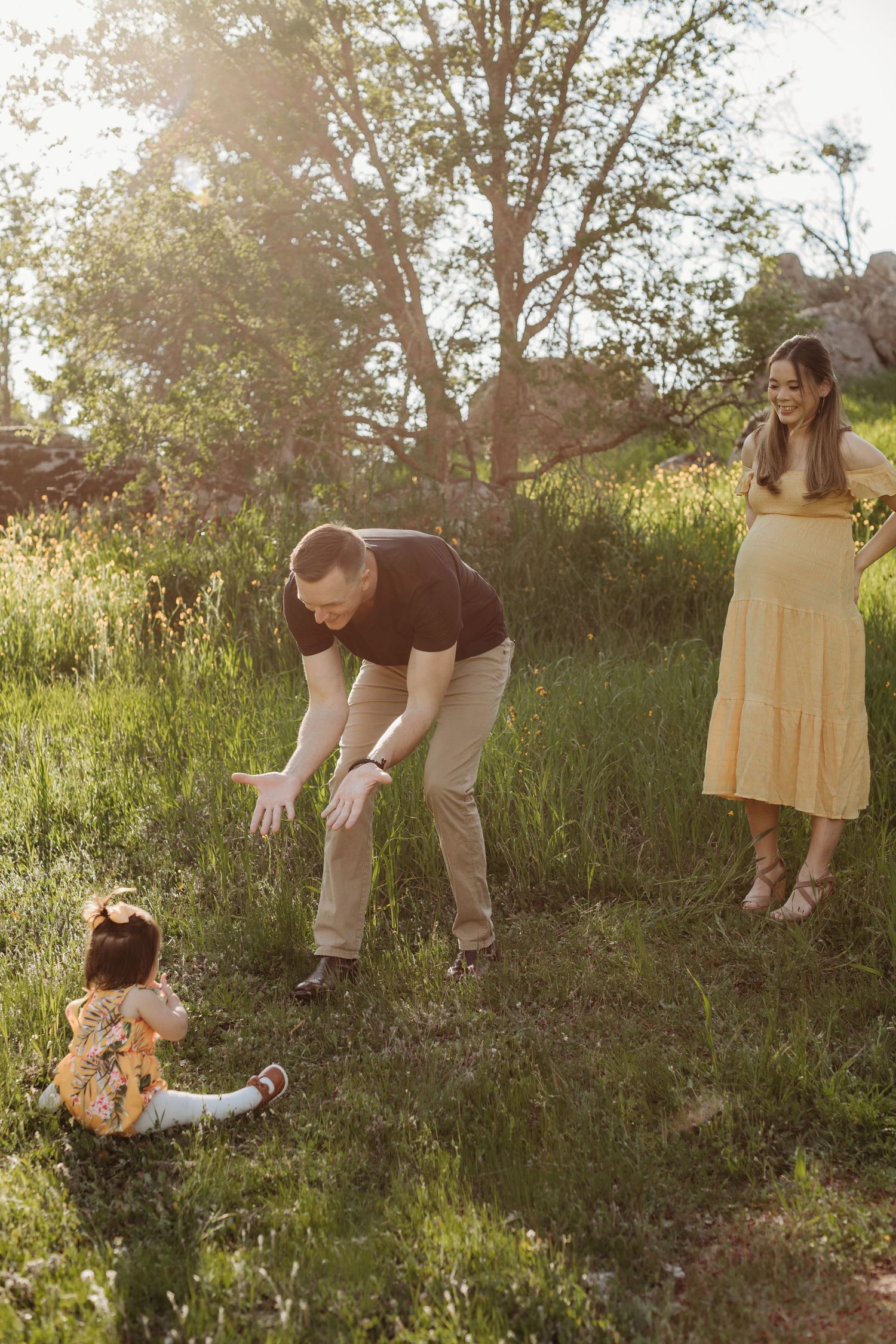 A child sits in grass while adults interact in a sunny outdoor setting.