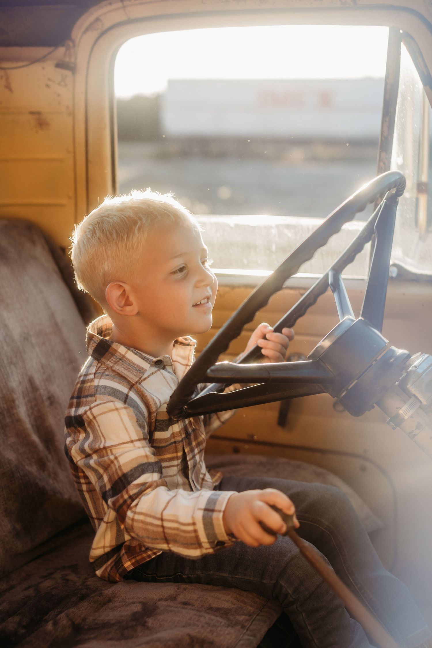 A child sits in the driver's seat of a vintage yellow vehicle, playfully gripping the steering wheel.