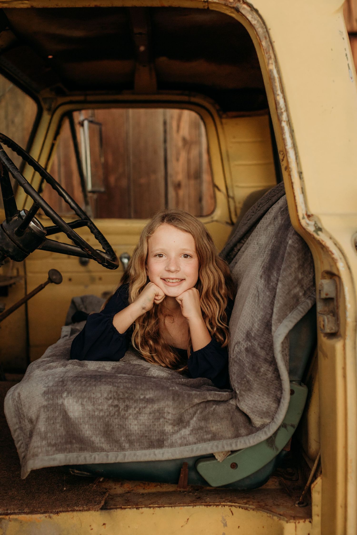Young child relaxing inside vintage yellow truck cab with cozy blanket.