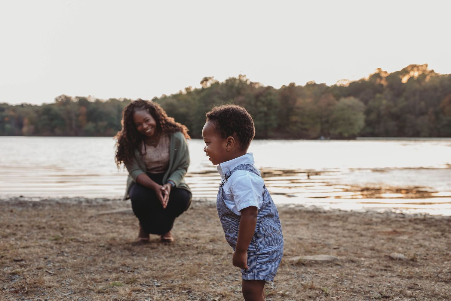 Mother and young son sharing a joyful moment on a sandy lakeside beach at sunset.
