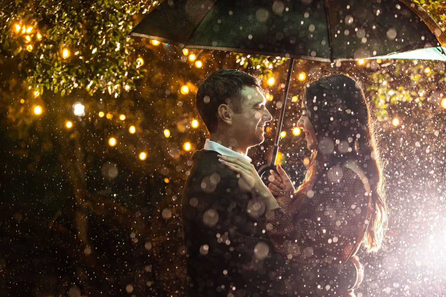 A young couple stands close together in the rain at night with string lights creating a magical romantic atmosphere.