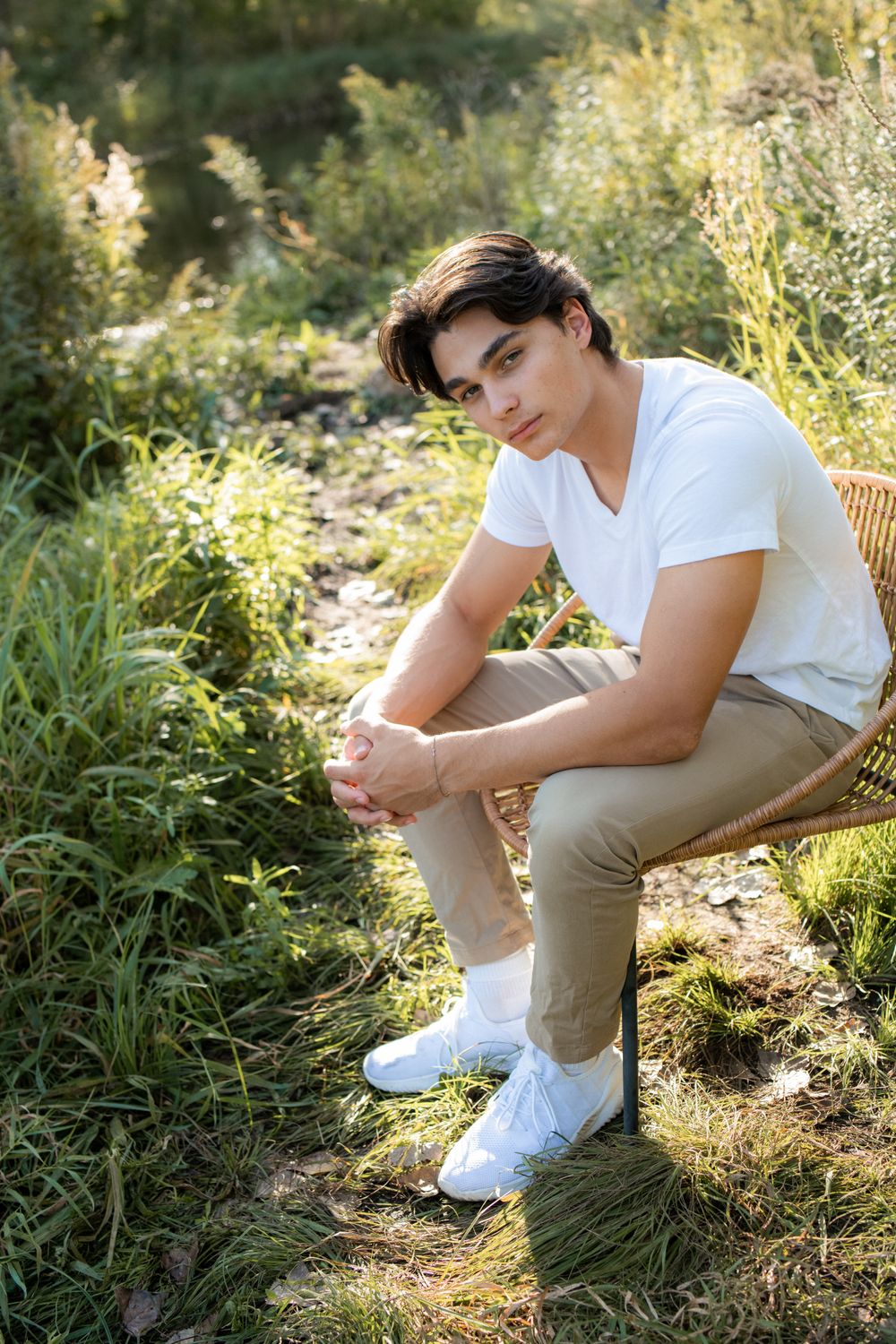 Casual outdoor senior portrait of person in white shirt sitting on wicker chair in natural setting.