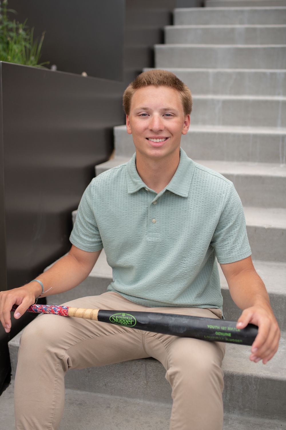A person in a mint green polo shirt and khaki pants sits on concrete steps holding a baseball bat.
