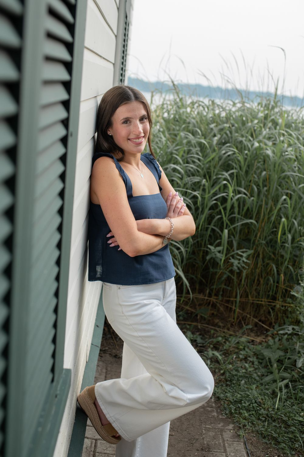 Woman in navy top and white pants poses outdoors near green plants and architectural columns.