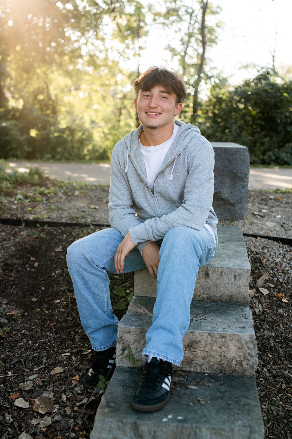 Person in a gray sweatshirt and jeans sits casually on stone steps during a sunny afternoon in an outdoor setting.